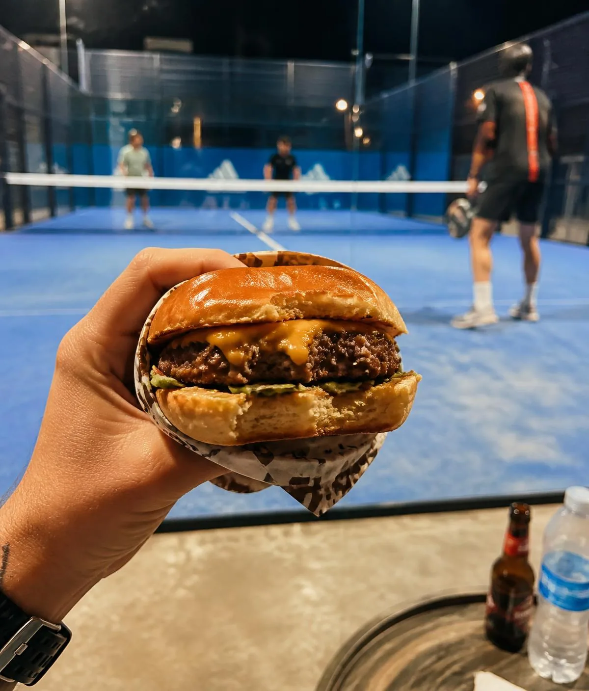 Hambúrguer artesanal da Smash Burger segurado em frente a uma quadra de padel